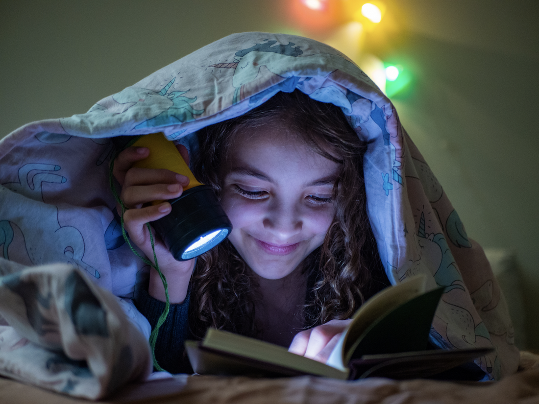 weighted blanket for ADHD: Girl reading a book in the dark using a flashlight while covered with a blanket