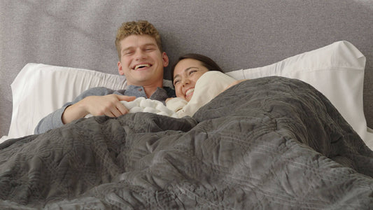 A couple smiles on a bed while snuggling under Hush sheets and blankets.