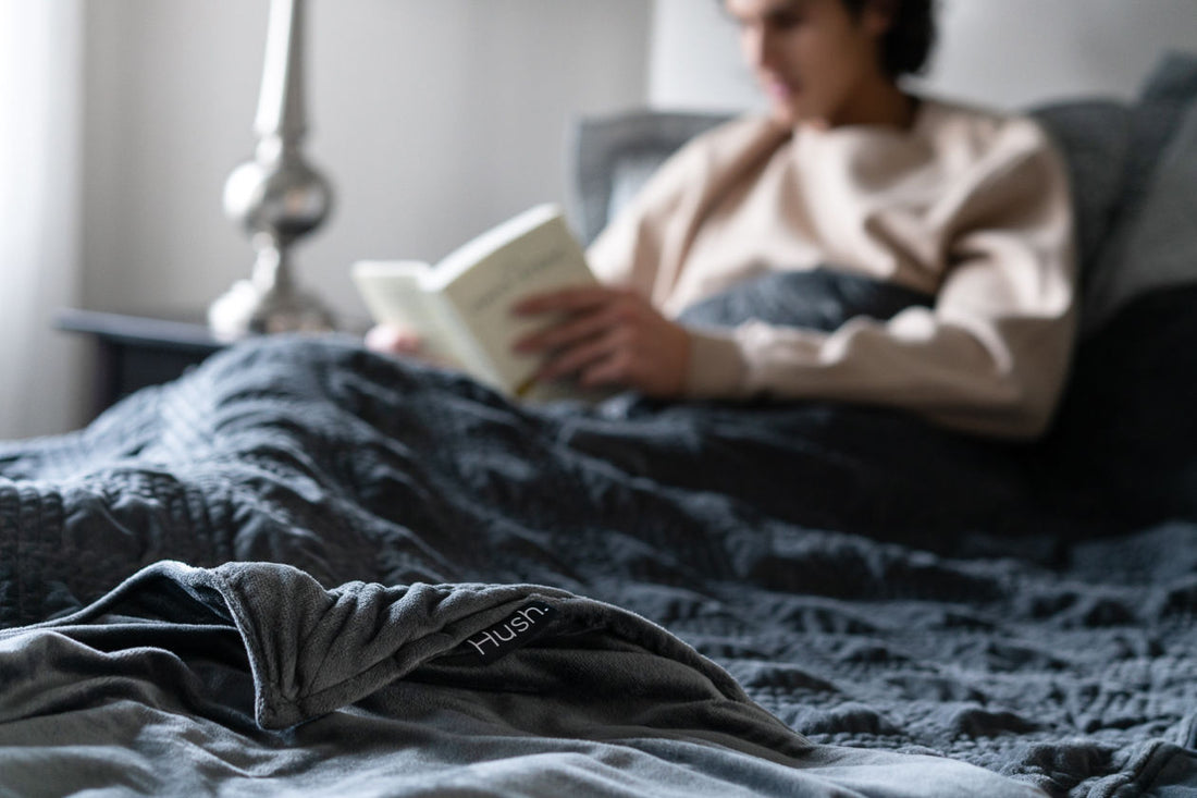 A man sitting in bed covered in grey Hush Classic Weighted Blanket while reading a book.