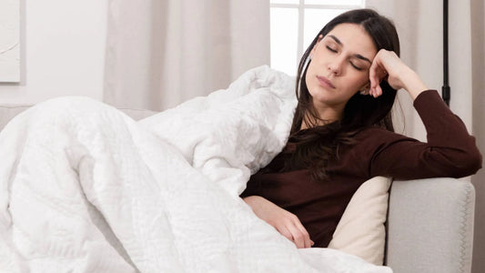 A young woman sleeps on the couch and under a white Hush Classic weighted blanket.