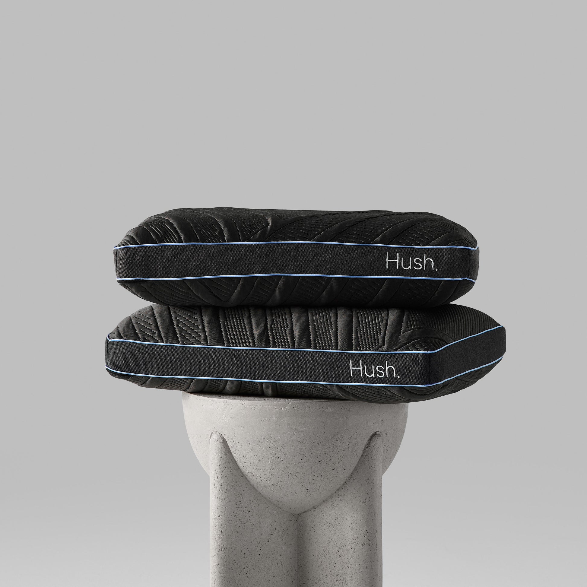 A black memory foam pillow with a blue accent on a textured grey base, labeled with the brand name 'HUSH'.